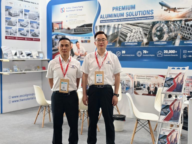 韩伟铝业团队在2026德国全球铝工业展展台前合影，背景为铝板、铝棒、锻环与铝管产品形象墙